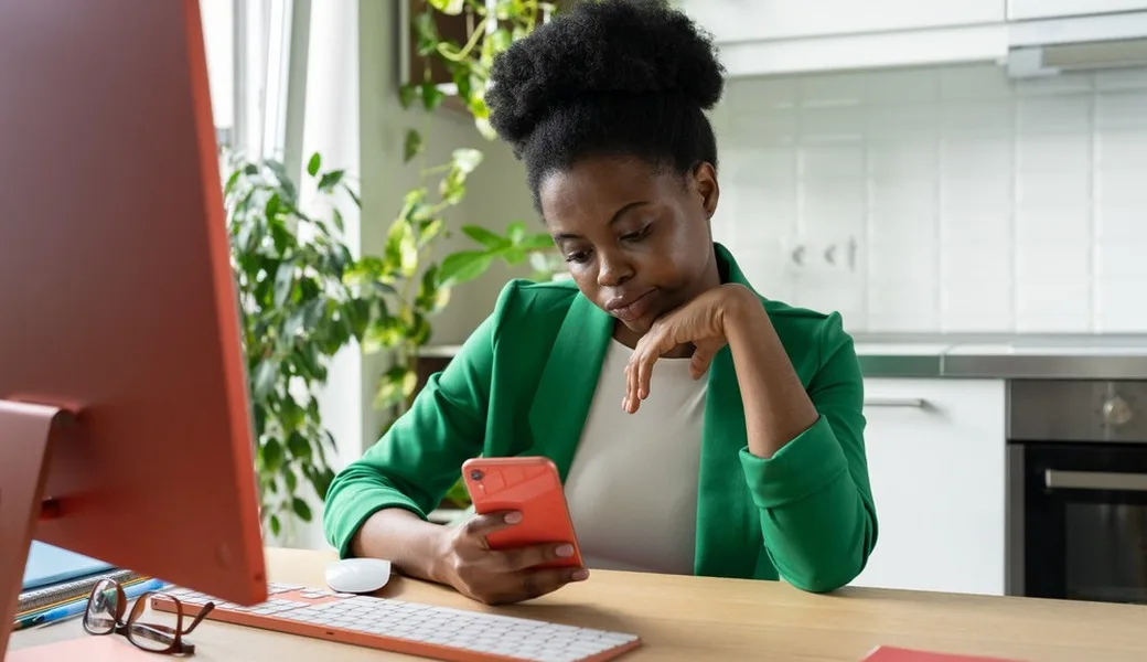 A woman procrastinating on her phone while sitting in front of a computer.
